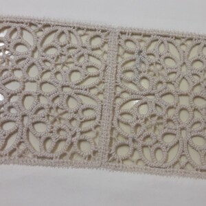 May include: A close-up of a beige crocheted lace trim with a repeating pattern of floral designs.
