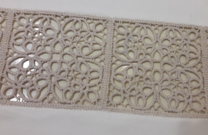 May include: A close-up of a beige crocheted lace trim with a repeating pattern of floral designs.
