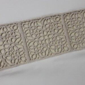 May include: A close-up of a white fabric with a beige lace trim. The lace trim features a repeating pattern of intricate floral designs.
