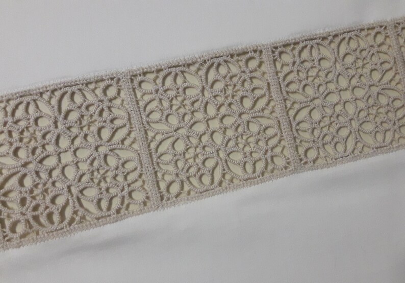 May include: A close-up of a white fabric with a beige lace trim. The lace trim features a repeating pattern of intricate floral designs.