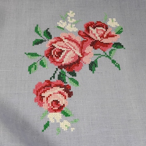 May include: A close-up of a cross-stitch embroidery design featuring three pink and red roses with green leaves and white flowers on a light gray linen fabric.