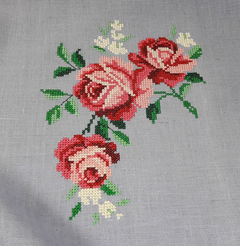 May include: A close-up of a cross-stitch embroidery design featuring three pink and red roses with green leaves and white flowers on a light gray linen fabric.