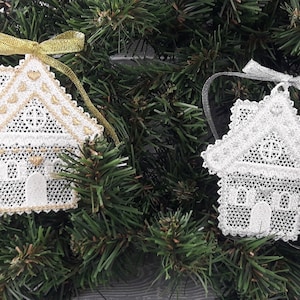 May include: Two white lace house ornaments with gold and silver ribbon. The ornaments are hanging on a green Christmas tree.