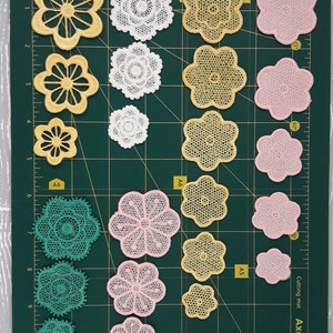 May include: Assortment of 15 delicate lace flower appliques in various colors, including white, yellow, pink, and green. The appliques are arranged on a green cutting mat with inch markings.