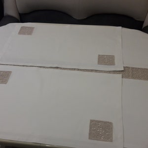 May include: Three white placemats with crocheted square designs in the centre. The placemats are on a white tablecloth with a crocheted border.