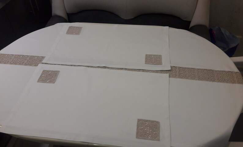 May include: Three white placemats with crocheted square designs in the centre. The placemats are on a white tablecloth with a crocheted border.