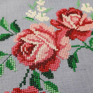 May include: Close-up of a cross-stitch embroidery design featuring pink and red roses with green leaves on a light gray fabric.