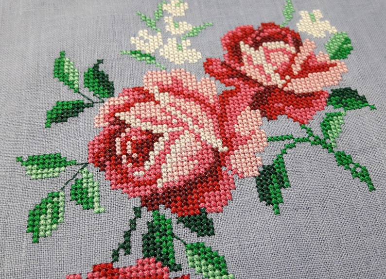 May include: Close-up of a cross-stitch embroidery design featuring pink and red roses with green leaves on a light gray fabric.