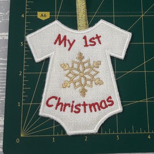 Pu&ograve; includere: Ciondolo in feltro bianco a forma di body per neonato con ricamo rosso che dice "Il mio 1&deg; Natale" e un fiocco di neve dorato al centro.