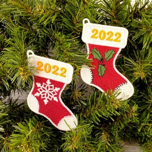 Puede incluir: Dos calcetines de Navidad de fieltro rojo con ribetes blancos, cada uno con un diseño diferente. Un calcetín tiene un copo de nieve y el otro una rama de acebo. Ambos calcetines tienen el año 2022 bordado en hilo dorado.