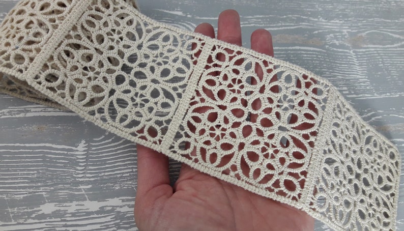 May include: A close-up of a cream-coloured lace trim featuring a repeating pattern of intricate floral designs. The lace is delicate and has a soft, airy feel.