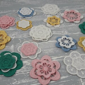 May include: Assortment of layered lace flower appliques in various colors, including pink, yellow, green, and blue. The appliques are arranged on a light gray wooden surface.