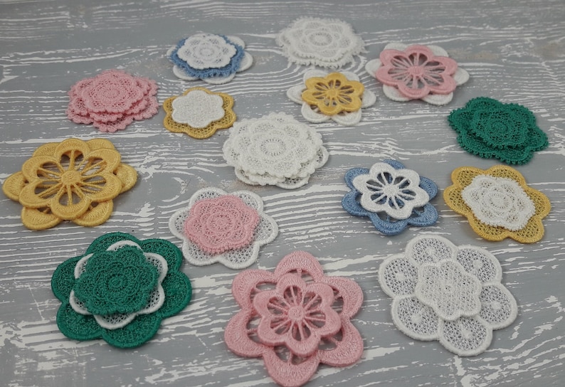 May include: Assortment of layered lace flower appliques in various colors, including pink, yellow, green, and blue. The appliques are arranged on a light gray wooden surface.