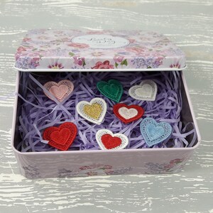 May include: A pink floral tin box with a lid that says "Lucky Day" is open and filled with purple shredded paper.  There are eight embroidered felt hearts in various colors, including red, pink, green, blue, and gold, inside the box.