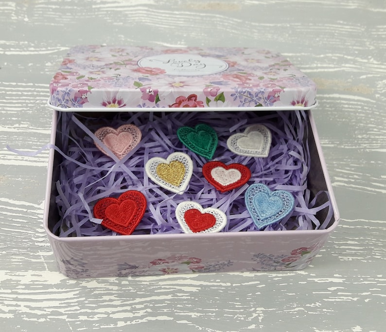 May include: A pink floral tin box with a lid that says "Lucky Day" is open and filled with purple shredded paper.  There are eight embroidered felt hearts in various colors, including red, pink, green, blue, and gold, inside the box.