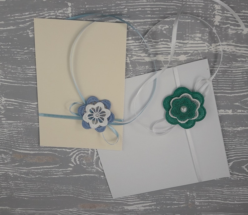May include: Two white cards with blue and green ribbon tied around them. The ribbon is tied in a bow with a blue and white flower on one card and a green flower on the other.
