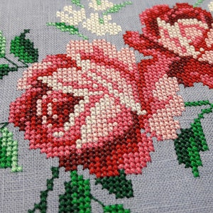 May include: Close-up of a cross-stitch embroidery featuring two pink and red roses with green leaves on a light gray fabric.