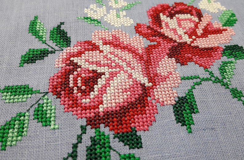 May include: Close-up of a cross-stitch embroidery featuring two pink and red roses with green leaves on a light gray fabric.