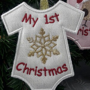 Pu&ograve; includere: Ciondolo in tessuto bianco a forma di body per neonato con un fiocco di neve dorato e ricamo rosso che recita "My 1st Christmas".