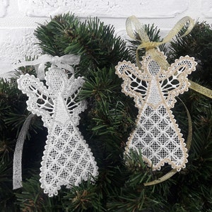 以下が含まれることがあります： 金と銀のリボンが付いた白いレースの天使のオーナメント2つ。天使は緑色の松の枝にぶら下がっています。