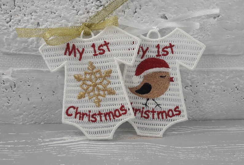Pu&ograve; includere: Due body da beb&eacute; in crochet bianco con ricami rossi. Un body ha un fiocco di neve e il testo "My 1st Christmas". L'altro body ha un pettirosso con berretto rosso e il testo "My 1st Christmas".