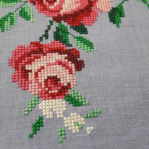 May include: Close-up of a cross-stitch embroidery featuring a cluster of pink and red roses with green leaves on a light gray linen fabric.