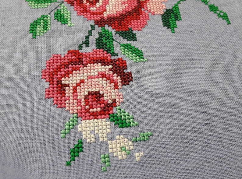 May include: Close-up of a cross-stitch embroidery featuring a cluster of pink and red roses with green leaves on a light gray linen fabric.