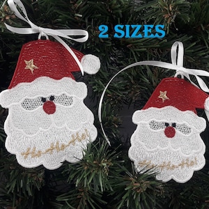 May include: Two white embroidered Santa Claus ornaments with red hats and gold trim, hanging on a green Christmas tree with white ribbon.