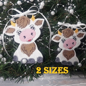 以下が含まれることがあります： 白い毛並み、茶色の斑点、ピンクの鼻を持つ、刺繍された牛のオーナメントが2つ。1つは他方より大きい。オーナメントは、白いリボンで緑の松の枝に、白い真珠のガーランドと一緒に吊るされています。
