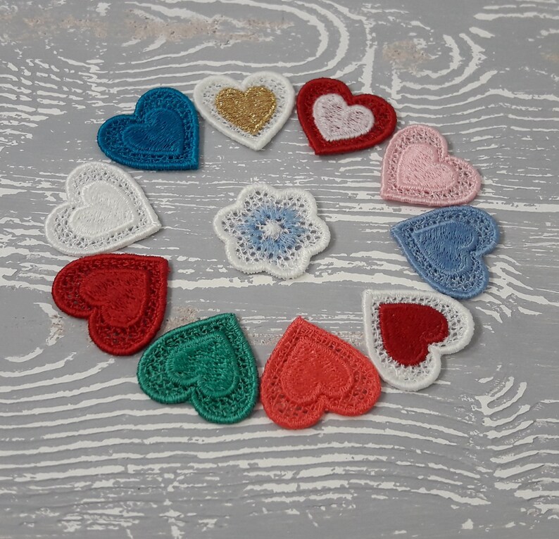 May include: A collection of ten embroidered fabric hearts in various colors, including red, pink, blue, green, and white. The hearts are arranged in a circle on a white wooden surface.