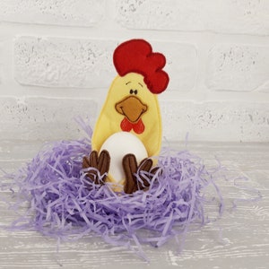 May include: A yellow felt chicken with a red comb and brown feet holds a white egg in a purple shredded paper nest.