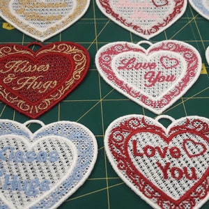 Pode incluir: Nove ornamentos bordados em forma de cora&ccedil;&atilde;o com v&aacute;rias mensagens do Dia dos Namorados, incluindo "Love You", "For You" e "Kisses & Hugs". Os ornamentos s&atilde;o brancos com bordados vermelhos, rosa, azuis e dourados.