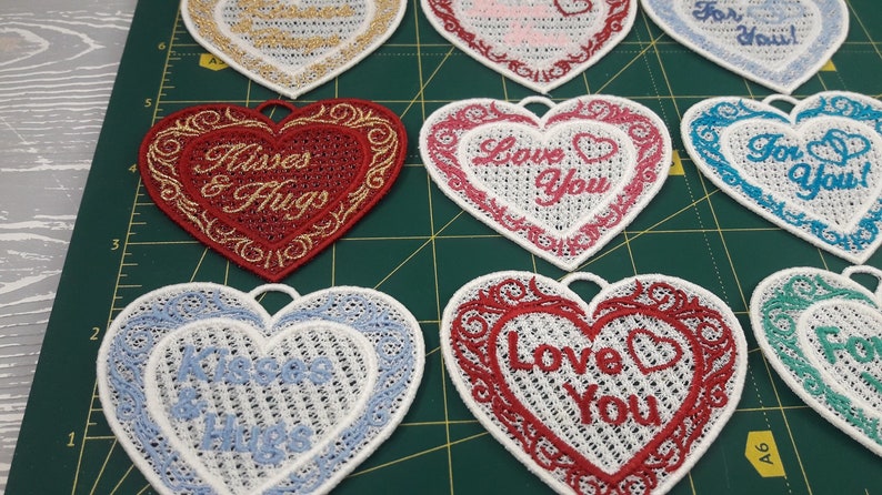 Pode incluir: Nove ornamentos bordados em forma de cora&ccedil;&atilde;o com v&aacute;rias mensagens do Dia dos Namorados, incluindo "Love You", "For You" e "Kisses & Hugs". Os ornamentos s&atilde;o brancos com bordados vermelhos, rosa, azuis e dourados.