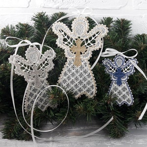 以下が含まれることがあります： 繊細なレースのディテールが施された、3つの装飾的な天使のオーナメント。1つはシルバー、1つは金の十字架が付いた白、3つ目は青い十字架が付いた白です。それぞれに白いリボンが付いており、松の枝を背景に飾られています。