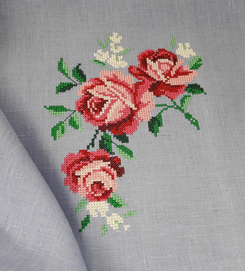 May include: A close-up of a gray linen fabric with a cross-stitch embroidery design of pink and red roses with green leaves.