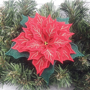May include: A red poinsettia flower with gold accents, embroidered on a green fabric. The flower is surrounded by green pine branches.