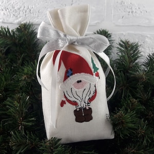 May include: A white linen gift bag with a silver ribbon and an embroidered gnome design. The gnome is wearing a red hat, a white beard, and brown boots.