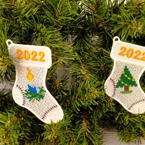 Puede incluir: Dos calcetines de Navidad de fieltro blanco con el año 2022 bordado en la parte superior. Un calcetín tiene un árbol de Navidad verde con luces amarillas bordadas. El otro calcetín tiene una vela azul con una llama amarilla bordada.