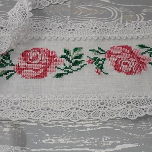 May include: White rectangular fabric with a cross-stitched floral design featuring pink roses and green leaves. The edges are trimmed with delicate white lace, creating a vintage or handmade aesthetic.