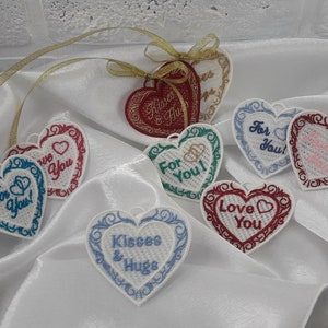 Pode incluir: Oito ornamentos brancos em forma de cora&ccedil;&atilde;o com mensagens bordadas. As mensagens incluem "Para voc&ecirc;!" "Amo voc&ecirc;" "Beijos e abra&ccedil;os" "Para voc&ecirc;" e "Amamos voc&ecirc;".