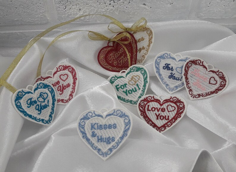 Pode incluir: Oito ornamentos brancos em forma de cora&ccedil;&atilde;o com mensagens bordadas. As mensagens incluem "Para voc&ecirc;!" "Amo voc&ecirc;" "Beijos e abra&ccedil;os" "Para voc&ecirc;" e "Amamos voc&ecirc;".