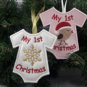 Pu&ograve; includere: Due ornamenti in tessuto bianco a forma di body per neonato con ricami rossi. Un ornamento ha un fiocco di neve dorato e il testo "My 1st Christmas". L'altro ornamento ha un uccello rosso e marrone con un cappello di Babbo Natale e il testo "My 1st Christmas".