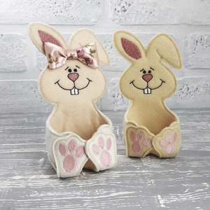 Può includere: Due cestini pasquali a forma di coniglio in feltro con orecchie rosa e impronte di zampe. Un coniglio ha un fiocco rosa con stampa floreale.