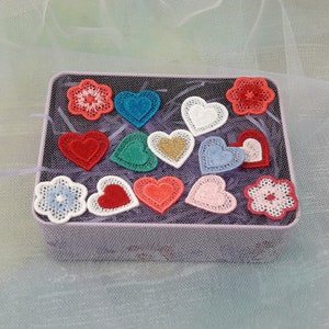 May include: A tin box filled with various embroidered patches in the shape of hearts and flowers. The patches are in shades of red, pink, blue, green, and white.