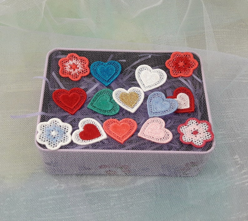 May include: A tin box filled with various embroidered patches in the shape of hearts and flowers. The patches are in shades of red, pink, blue, green, and white.