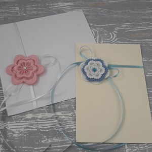 May include: Two white envelopes with decorative flowers. The envelope on the left has a pink flower with a white ribbon. The envelope on the right has a blue flower with a blue ribbon.