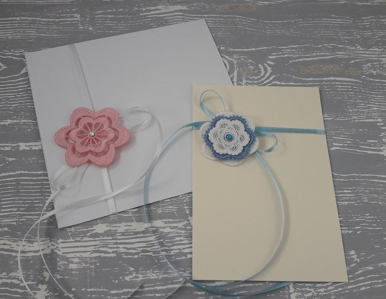 May include: Two white envelopes with decorative flowers. The envelope on the left has a pink flower with a white ribbon. The envelope on the right has a blue flower with a blue ribbon.