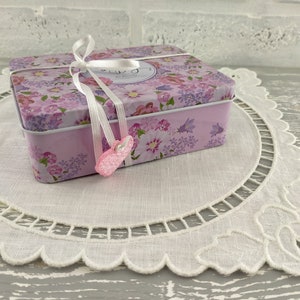 May include: A pink floral tin box with a white ribbon and a pink heart charm. The box is sitting on a white crocheted doily.