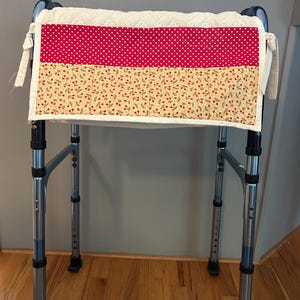 May include: A quilted walker bag with a white eyelet top and a red polka dot and cherry print design. The bag is attached to a silver walker frame, providing storage and a decorative touch.