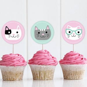 Puede incluir: Tres cupcakes rosas con glaseado blanco cubiertos con adornos redondos de papel para cupcakes que representan diferentes gatos de dibujos animados. Los adornos de gato están en palitos. El primer gato es blanco y negro, el segundo es gris y el tercero es blanco con gafas.